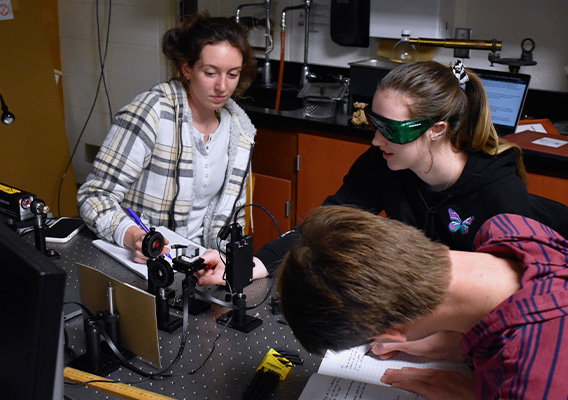 Two female student study an optical device while a third student takes notes in a physics lab classroom