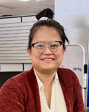 Photo of Hsakushee Zan, a woman wearing a red sweater with a white shirt. She has black hair in a bun on top of her head and is wearing black framed glasses.