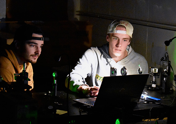 Two male students look at a laptop in a physics lab classroom