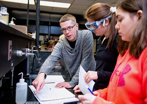 Chemistry Professor helps two students by pointing to a worksheet during a lab