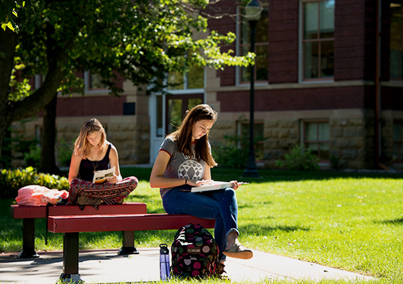 Two students sit on benches reading books in the sunshine